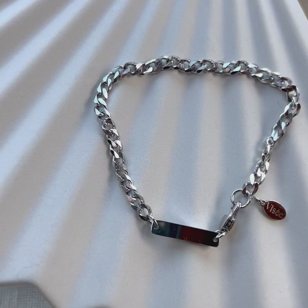Vidéo Bracelet "Libère toi" en acier argenté, style gourmette, avec plaque gravable, posé sur un fond beige texturé.