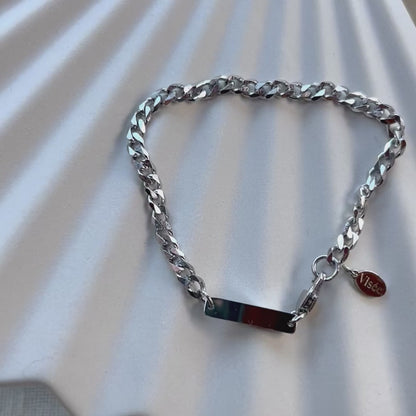 Vidéo Bracelet "Libère toi" en acier argenté, style gourmette, avec plaque gravable, posé sur un fond beige texturé.