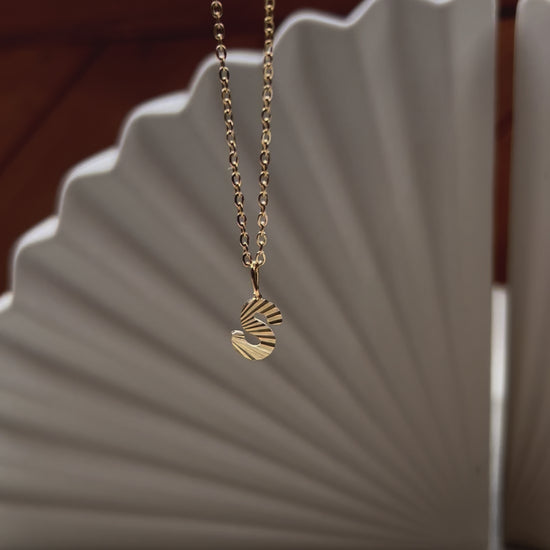 Vidéo du collier "Je suis" avec pendentif lettre striée, en acier doré, sur un fond beige texturé.