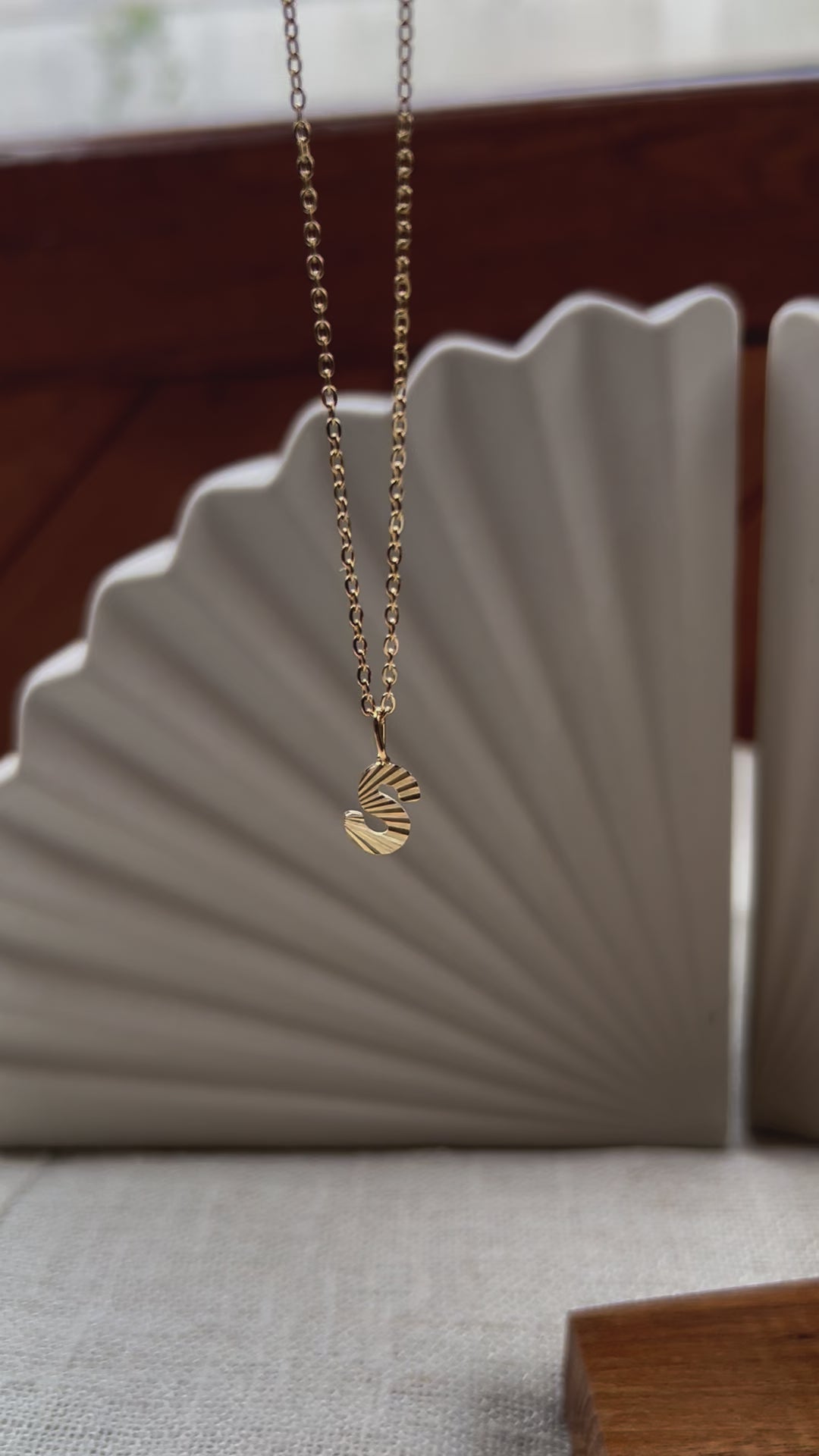 Vidéo du collier "Je suis" avec pendentif lettre striée, en acier doré, sur un fond beige texturé.