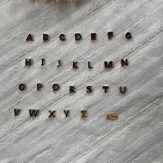 Vidéo de l'ensemble des pendentifs lettres dorées en acier inoxydable, soigneusement alignés  sur un tissu beige texturé.