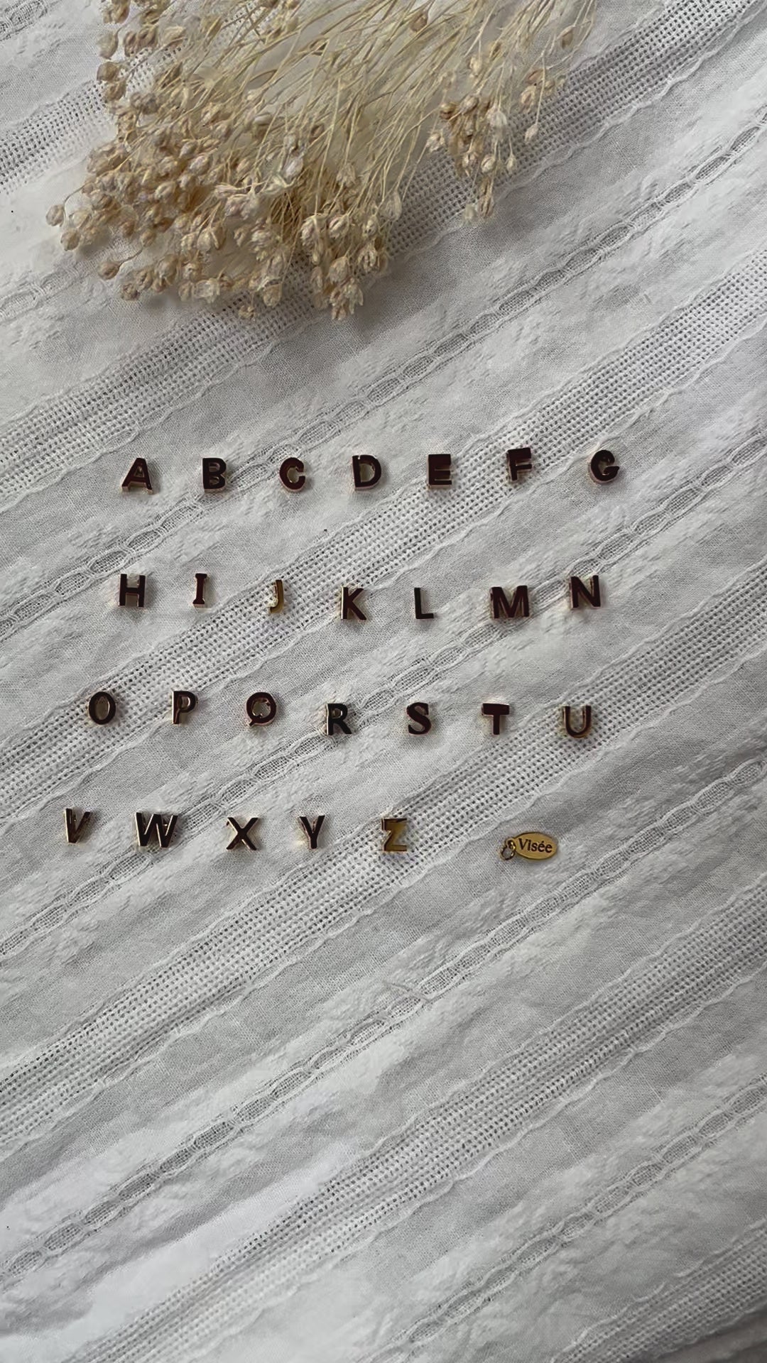 Vidéo de l'ensemble des pendentifs lettres dorées en acier inoxydable, soigneusement alignés  sur un tissu beige texturé.