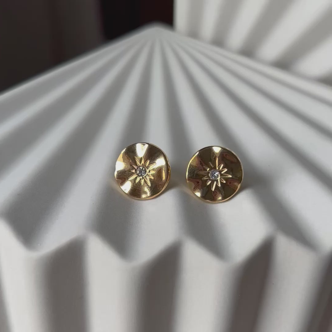 Vidéo des boucles d’oreilles Céleste dorées motif étoile, serties d'un zircon, posées sur un fond beige.