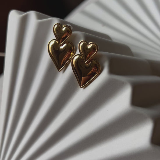 Vidéo des boucles d'oreilles "Elle m'a dit" en forme de double coeur, en acier. Posées sur un fond beige texturé. 