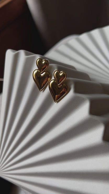 Vidéo des boucles d'oreilles "Elle m'a dit" en forme de double coeur, en acier. Posées sur un fond beige texturé. 