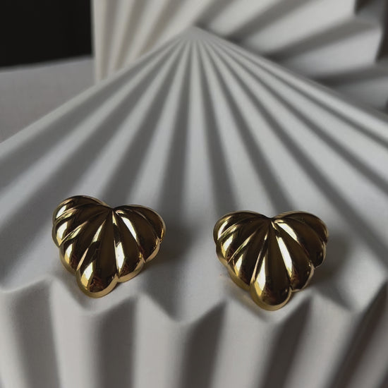 Vidéo des boucles d'oreilles "Coeur audacieux", en acier inoxydable, au design en relief en forme de coeur, posées côte à côte sur un fond beige texturé.