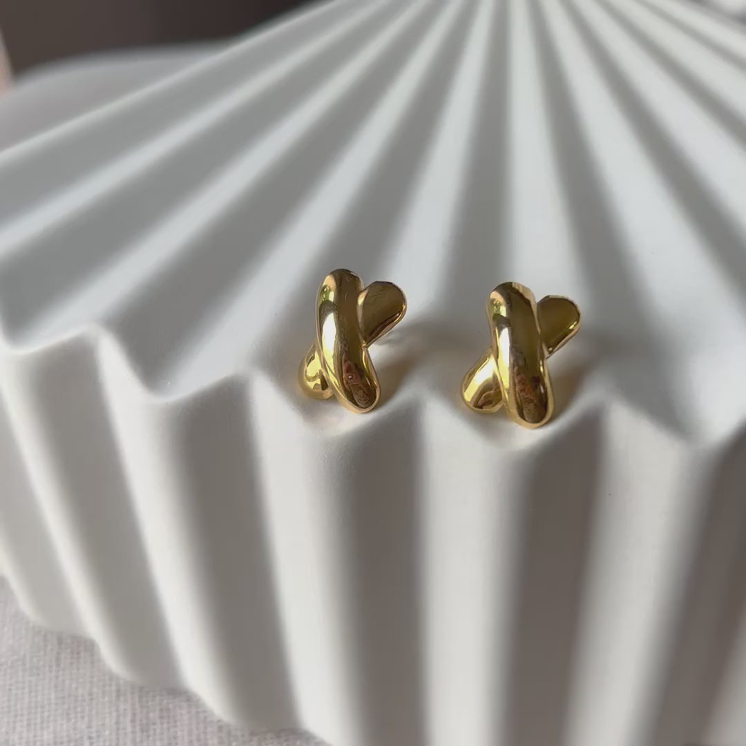 Vidéo des boucles d'oreilles "Révèle toi", au design croix en acier inoxydable doré, photographiées sur un fond beige texturé.