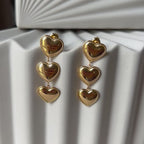 Vidéo boucles d'oreilles "Unique", en acier inoxydable doré, pendantes en cœurs, photographiées sur un fond beige texturé.