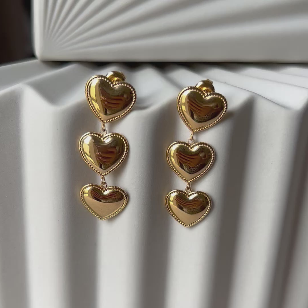 Vidéo boucles d'oreilles "Unique", en acier inoxydable doré, pendantes en cœurs, photographiées sur un fond beige texturé.