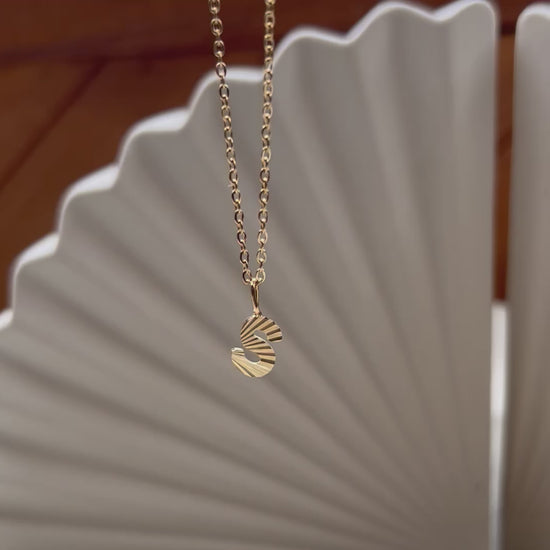 Vidéo du collier "Je suis" avec pendentif lettre striée, en acier doré, sur un fond beige texturé.