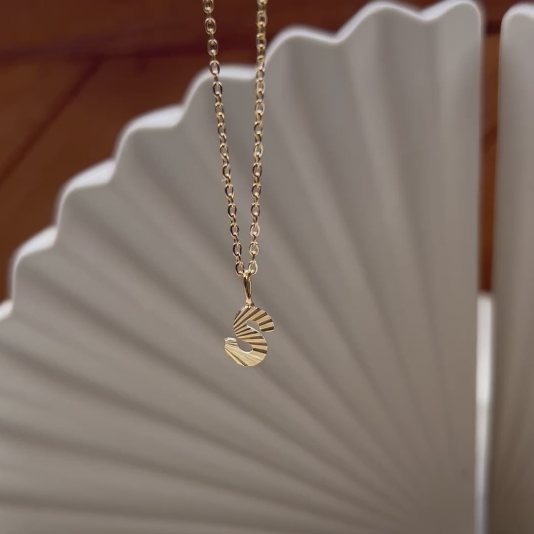 Vidéo du collier "Je suis" avec pendentif lettre striée, en acier doré, sur un fond beige texturé.