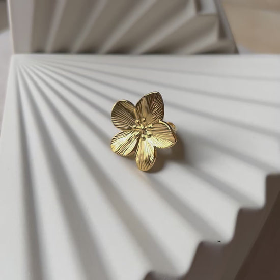 Vidéo bague réglable dorée en acier inoxydable, design fleur, vue rapprochée de face, posée sur un support beige texturé.