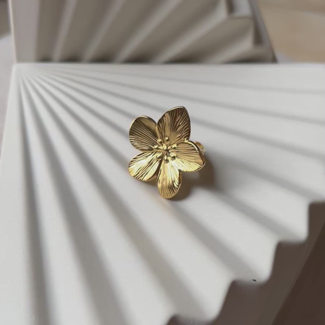 Vidéo bague réglable dorée en acier inoxydable, design fleur, vue rapprochée de face, posée sur un support beige texturé.