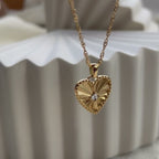 Vidéo sur le collier "Pensée" en acier doré avec pendentif cœur strié orné d'un zircon central, personnalisable, posé sur un fond beige texturé.