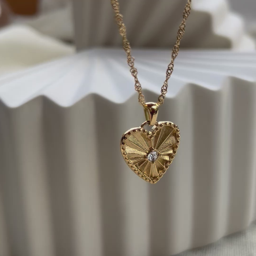 Vidéo sur le collier "Pensée" en acier doré avec pendentif cœur strié orné d'un zircon central, personnalisable, posé sur un fond beige texturé.