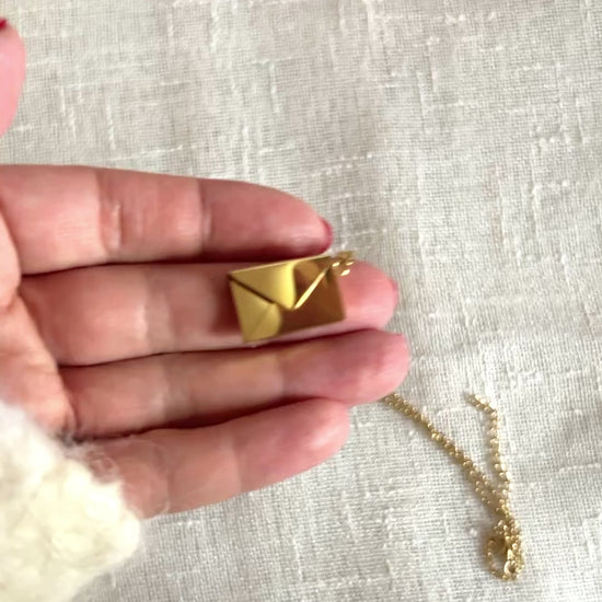 Vidéo du Collier "Nid secret", pendentif en forme d'enveloppe dorée ouverte, dévoilant un message, posé avec soin sur un fond beige texturé.
