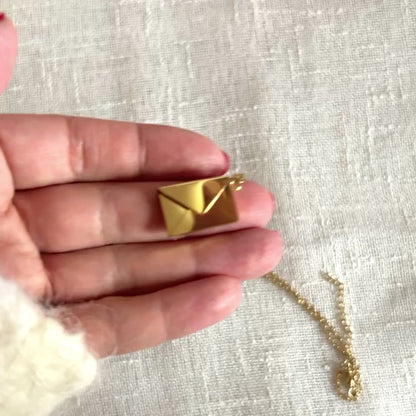 Vidéo du Collier "Nid secret", pendentif en forme d'enveloppe dorée ouverte, dévoilant un message, posé avec soin sur un fond beige texturé.