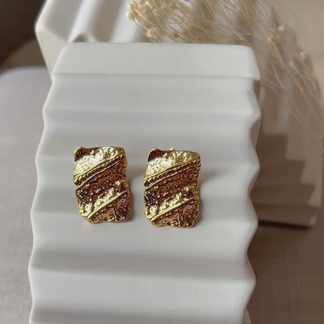 Vidéo boucles d'oreilles Affirmation rectangulaires texturées en acier doré, posées sur un fond beige texturé.