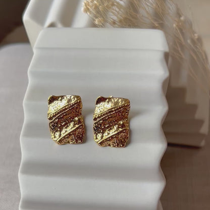 Vidéo boucles d'oreilles Affirmation rectangulaires texturées en acier doré, posées sur un fond beige texturé.
