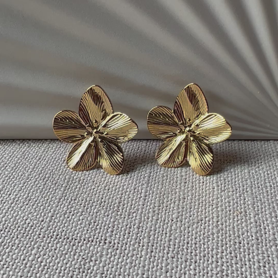 vidéo des boucles d'oreilles Fleur de peau en acier inoxydable dorée, motif floral oversize, posées sur un tissu beige texturé