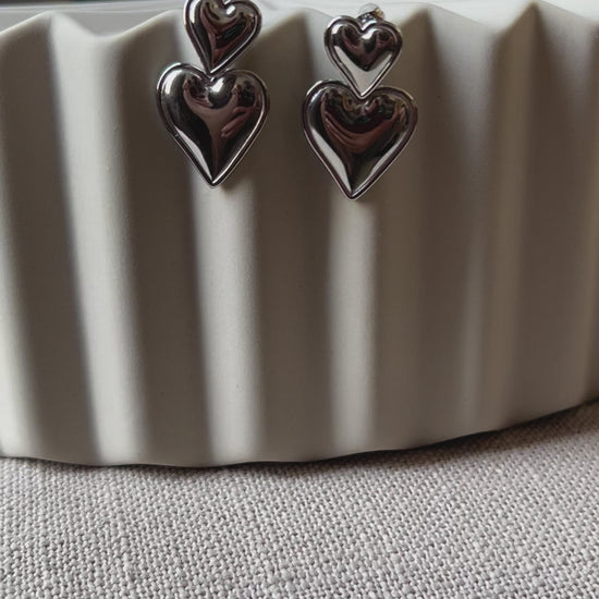 Vidéo des boucles d'oreilles "Elle m'a dit" en forme de double coeur, en acier. Posées sur un fond beige texturé. 