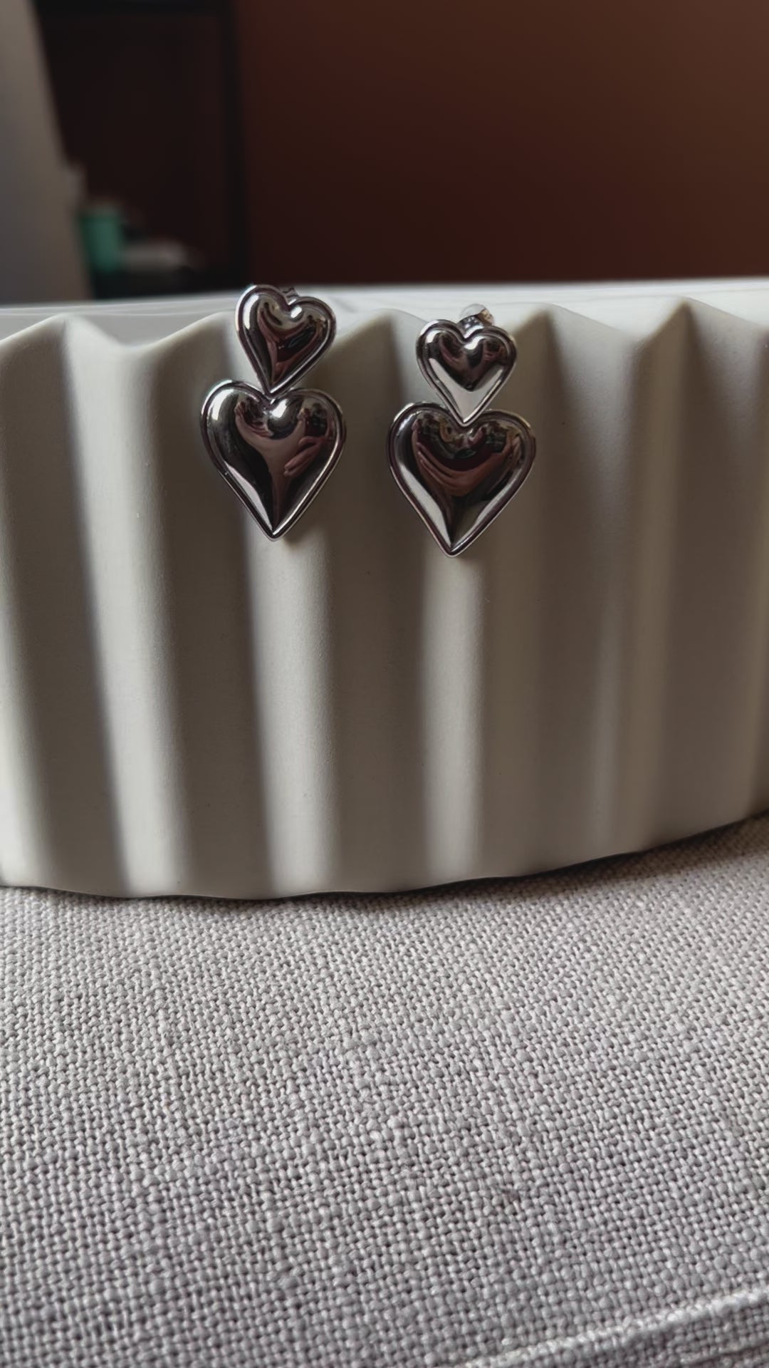 Vidéo des boucles d'oreilles "Elle m'a dit" en forme de double coeur, en acier. Posées sur un fond beige texturé. 