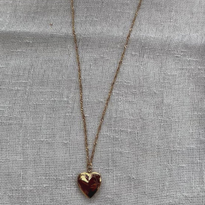 Vidéo du collier "Entre nous", médaillon cœur en acier doré avec photo souvenir à personnaliser, posé sur un fond beige texturé.