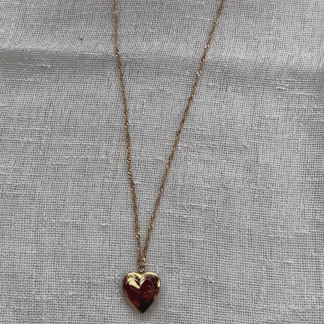 Vidéo du collier "Entre nous", médaillon cœur en acier doré avec photo souvenir à personnaliser, posé sur un fond beige texturé.