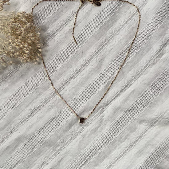 Vidéo du collier L'instant avec pendentif lettre E en acier inoxydable doré, posé sur un tissu beige doux.