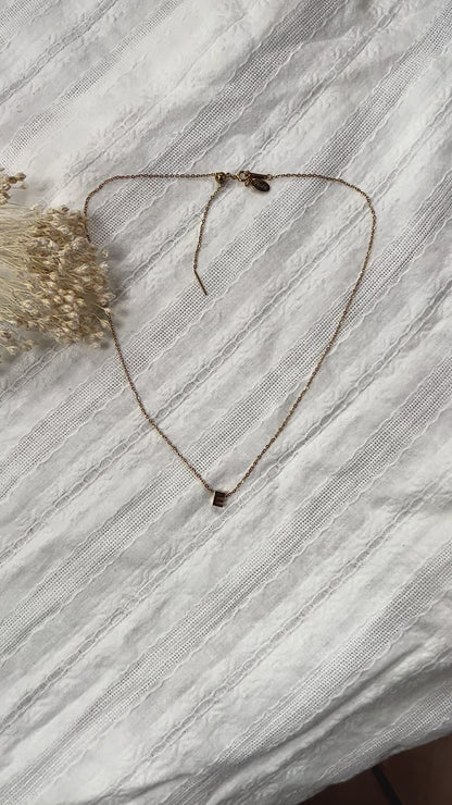 Vidéo du collier L'instant avec pendentif lettre E en acier inoxydable doré, posé sur un tissu beige doux.